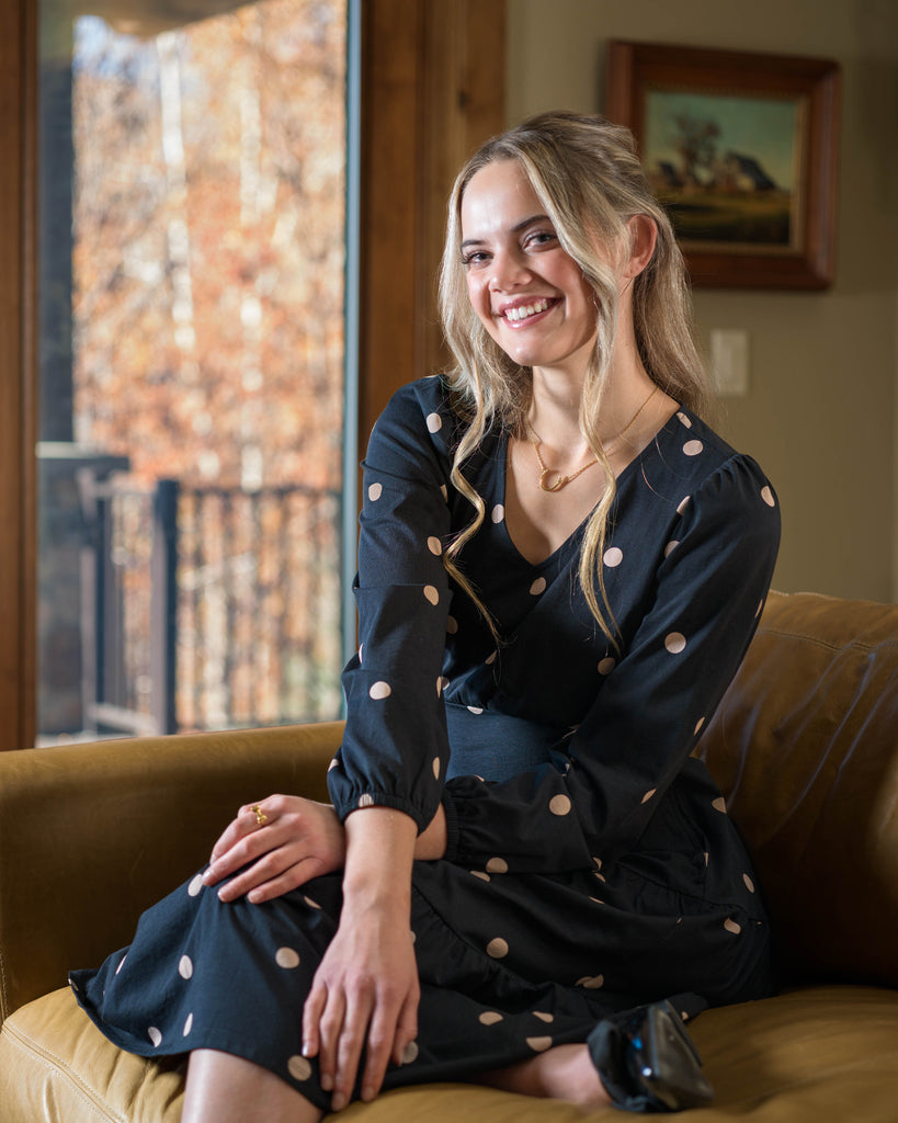 Model wearing a black with tan polka dot, v-neck, midi-length dress with tiers on skirt and pockets.