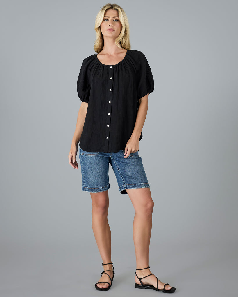 Model wearing a black, short sleeve, scoop neck blouse with white buttons down the front.