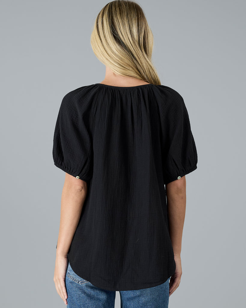 Model wearing a black, short sleeve, scoop neck blouse with white buttons down the front.