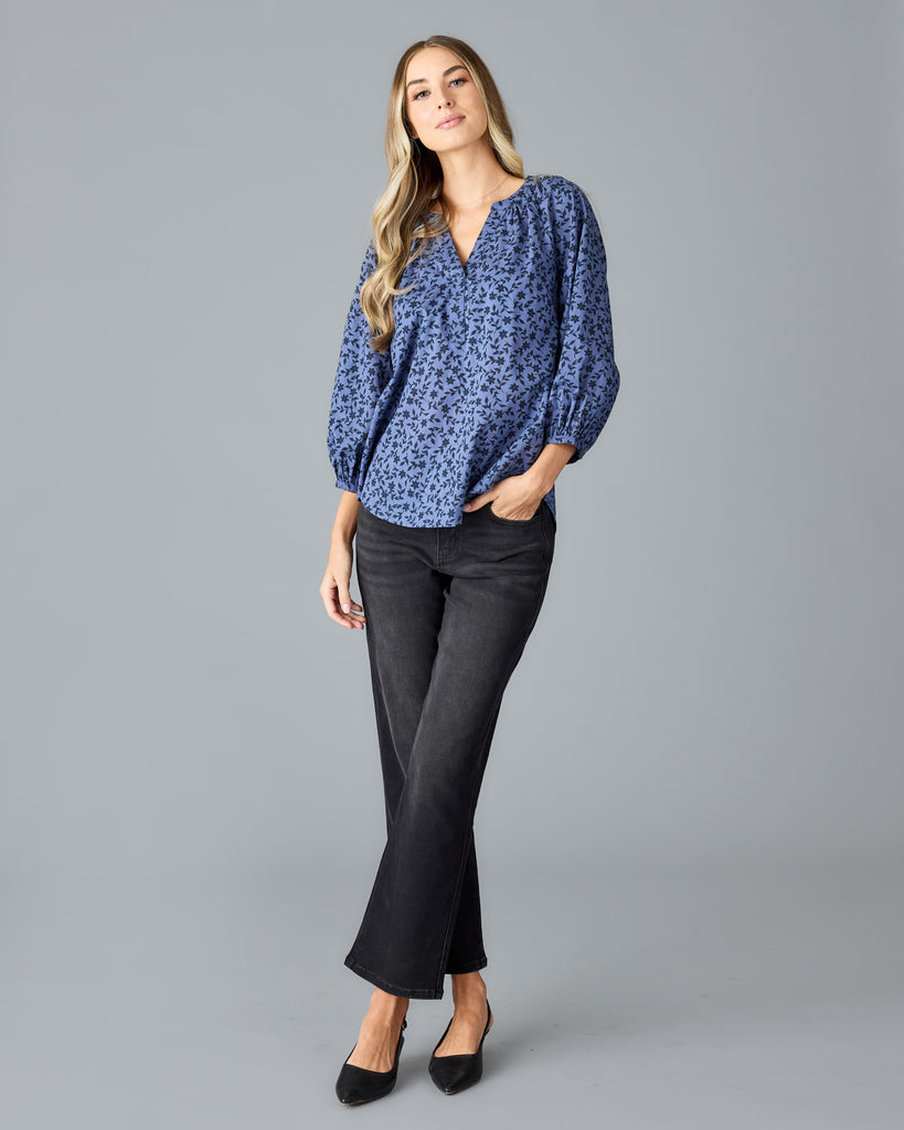 Model wearing a blue with black floral print long sleeve, v-neck, look fitting blouse.