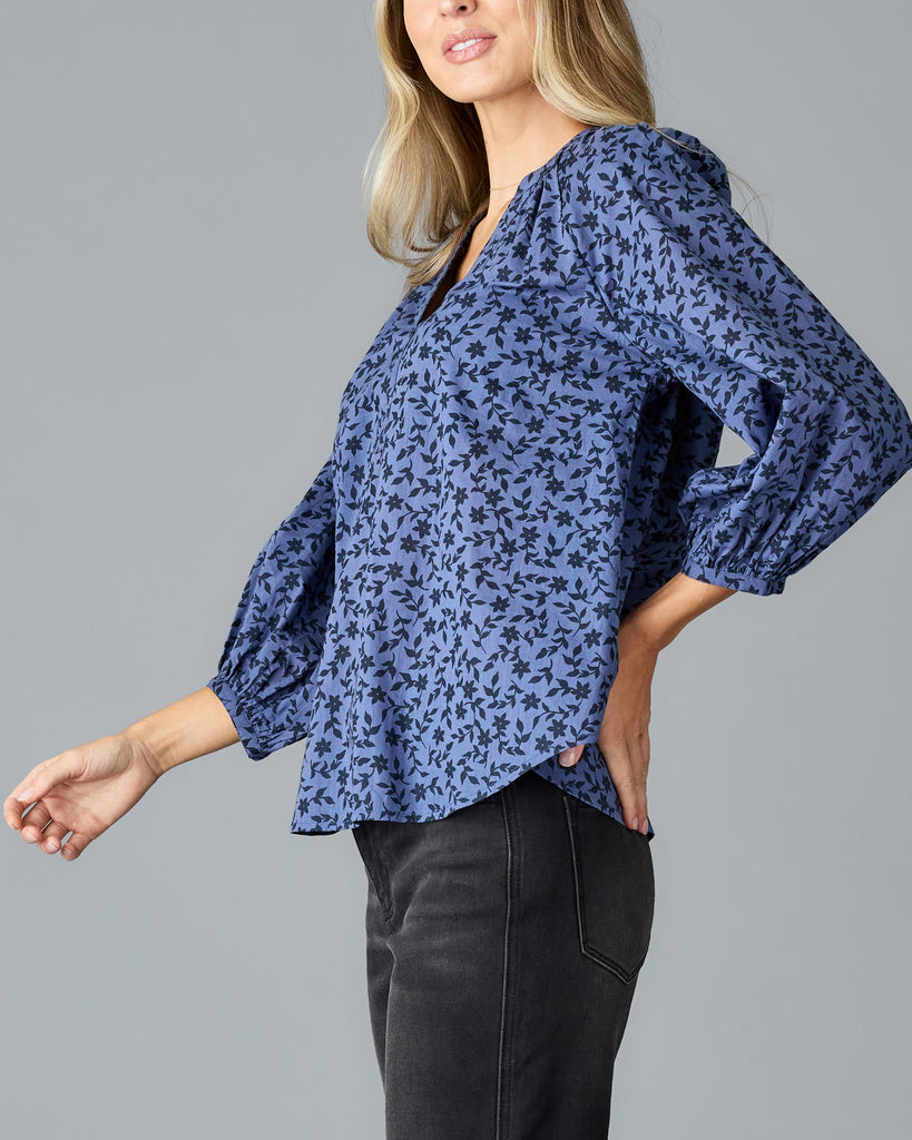 Model wearing a blue with black floral print long sleeve, v-neck, look fitting blouse.