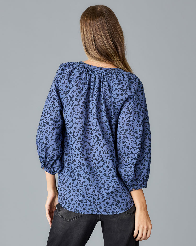 Model wearing a blue with black floral print long sleeve, v-neck, look fitting blouse.