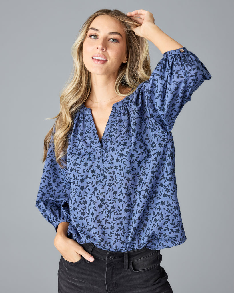 Model wearing a blue with black floral print long sleeve, v-neck, look fitting blouse.