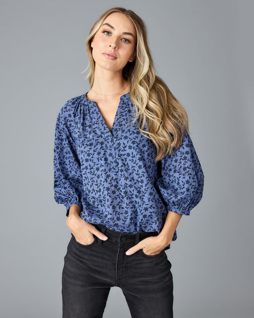 Model wearing a blue with black floral print long sleeve, v-neck, look fitting blouse.