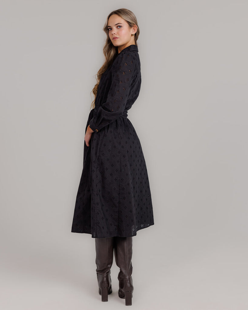 Model wearing a black long sleeve, collared, midi-length dress with lace fabric, buttons down the front and a sash at the waist.