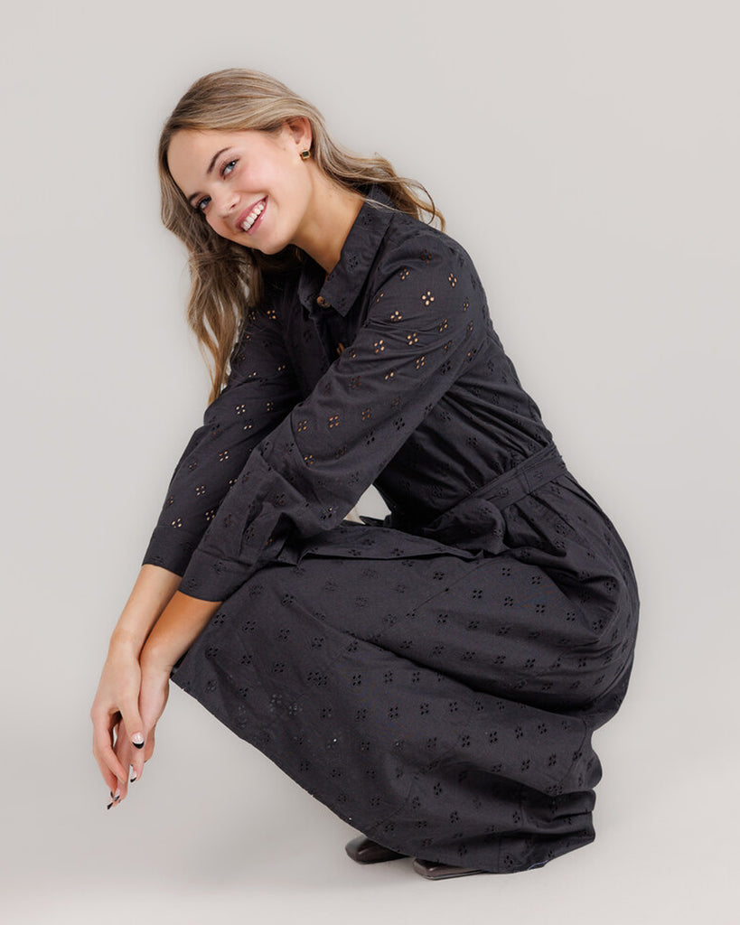 Model wearing a black long sleeve, collared, midi-length dress with lace fabric, buttons down the front and a sash at the waist.