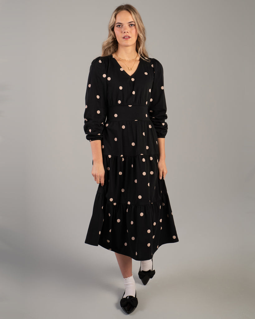 Model wearing a black with tan polka dot, v-neck, midi-length dress with tiers on skirt and pockets.