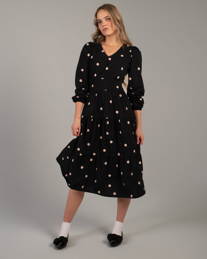 Model wearing a black with tan polka dot, knit, v-neck, midi-length dress with tiers on skirt and pockets.