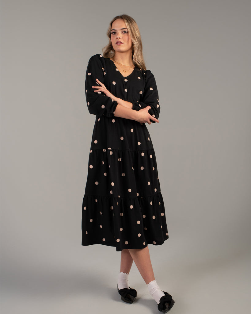 Model wearing a black with tan polka dot, v-neck, midi-length dress with tiers on skirt and pockets.