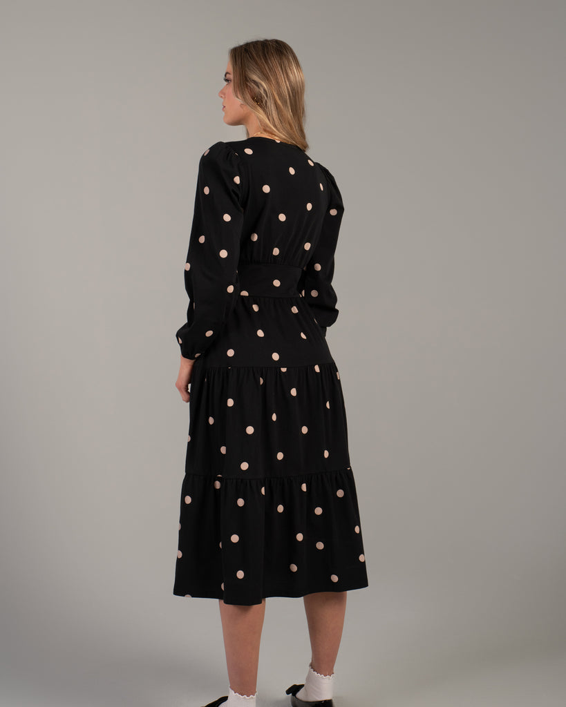 Back view of a model wearing a black with tan polka dot, midi-length dress with tiers on skirt and pockets.