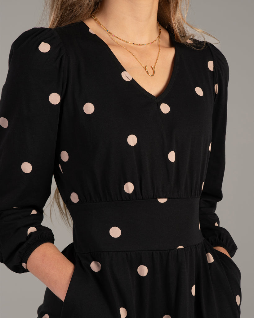 Close-up view of model wearing a black with tan polka dot, v-neck dress with pockets.