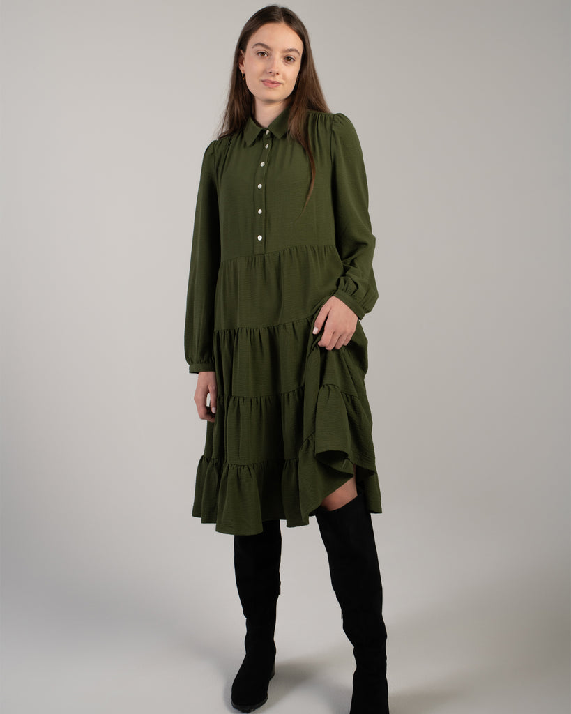 Model wearing a dark green long sleeve, knee-length dress with tiers and buttons down the front.