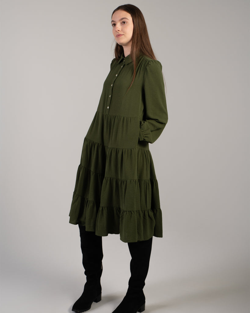 Model wearing a dark green long sleeve, knee-length dress with tiers and pockets.