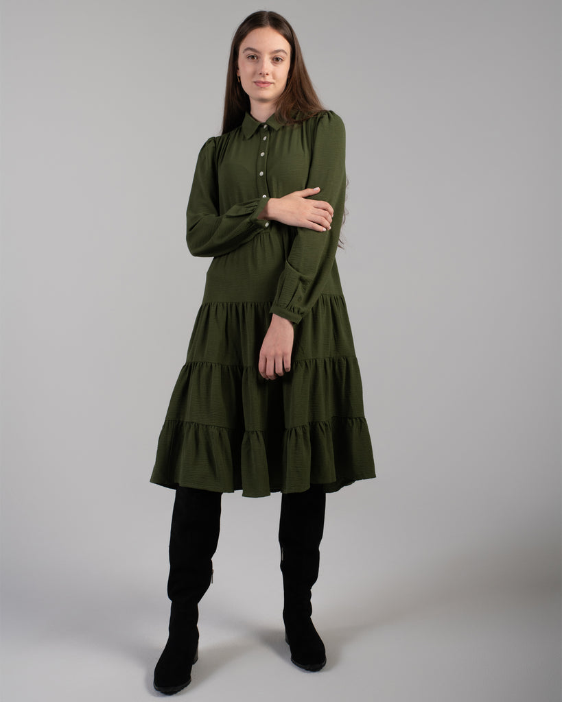 Model wearing a dark green long sleeve, knee-length dress with tiers and buttons down the front.