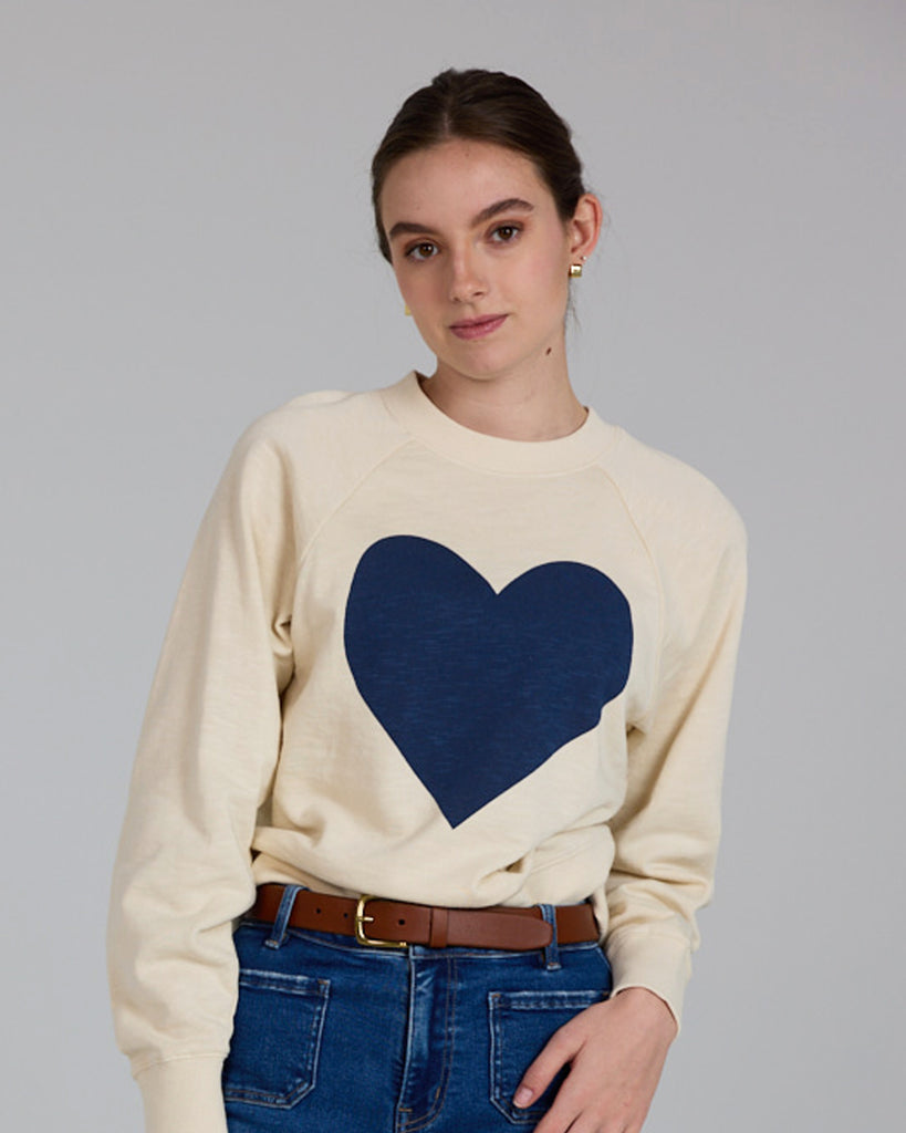 Model wearing an off-white with a blue heart print on the front, long sleeve crew neck sweatshirt.