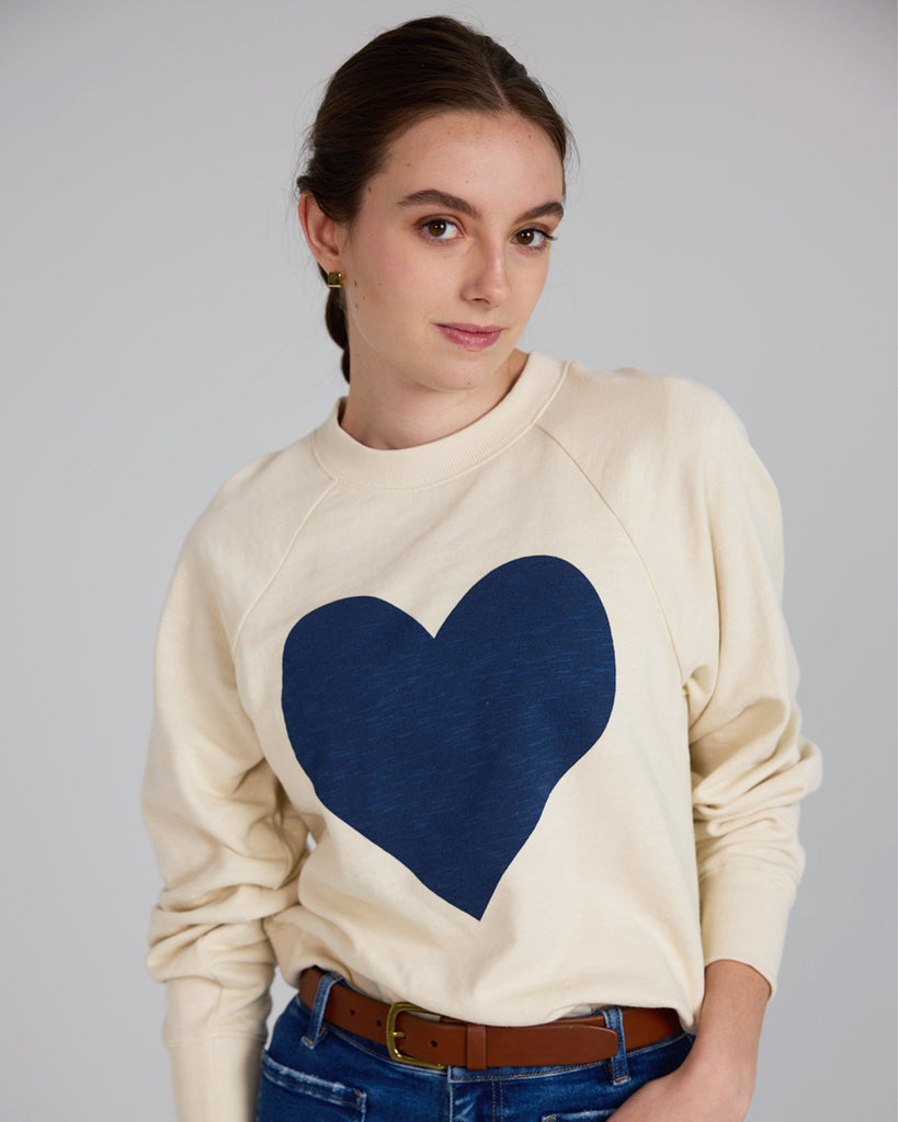 Model wearing an off-white with a blue heart print on the front, long sleeve crew neck sweatshirt.