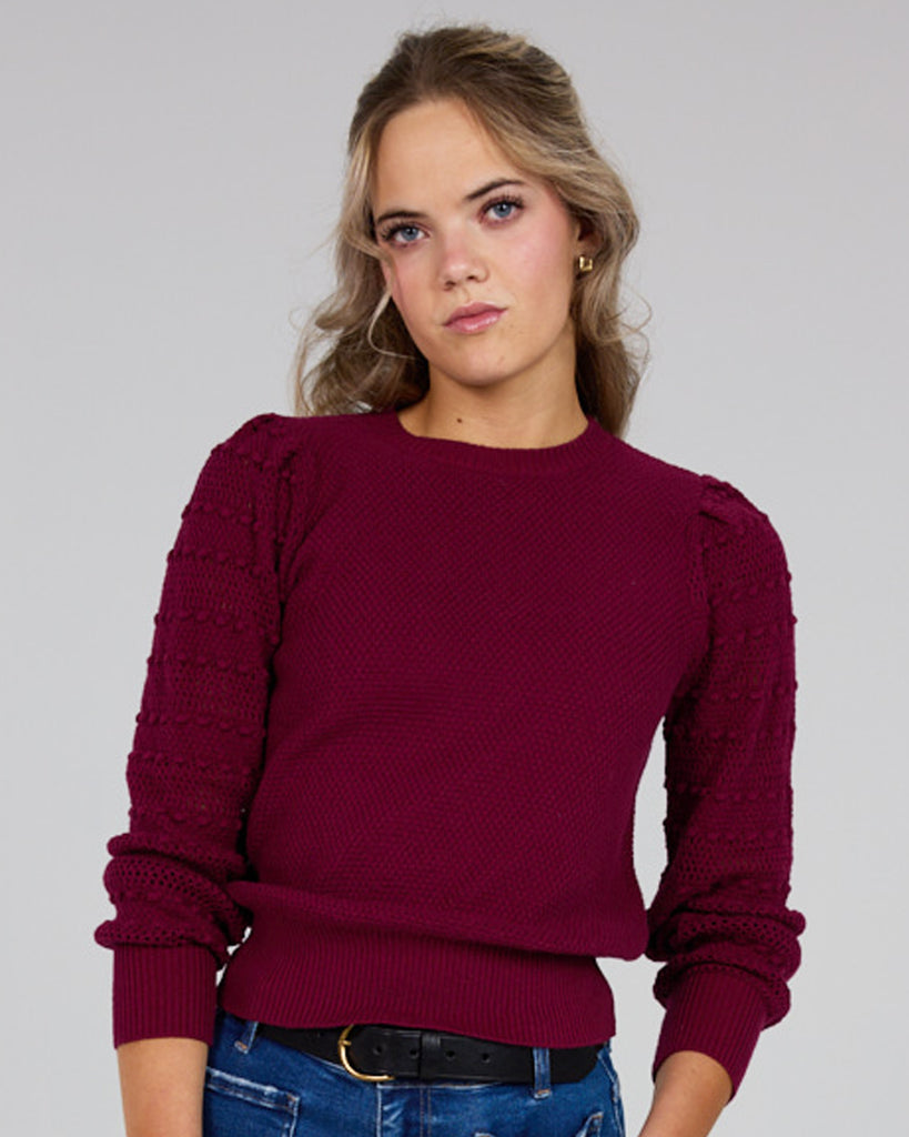 Model wearing a burgundy long sleeve sweater with pom pom details down sleeves.