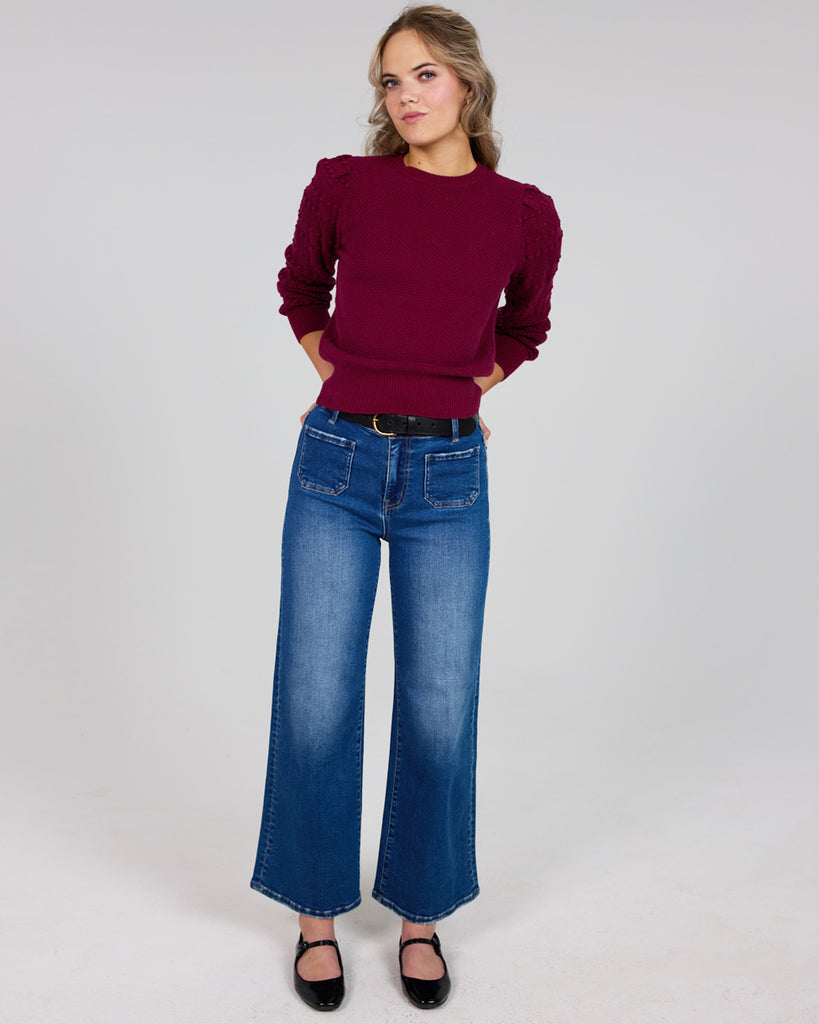 Model wearing a burgundy long sleeve sweater with pom pom details down sleeves and denim pants.