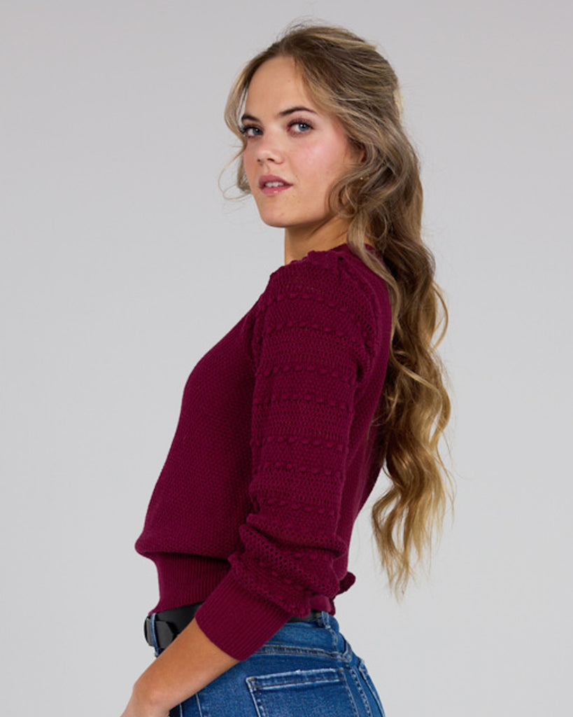 Model wearing a burgundy long sleeve sweater with pom pom details down sleeves.