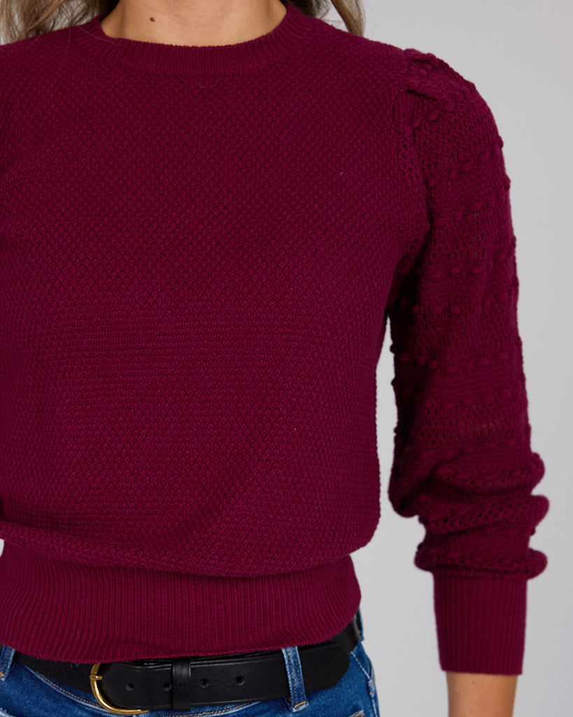 Model wearing a burgundy long sleeve sweater with pom pom details down sleeves.