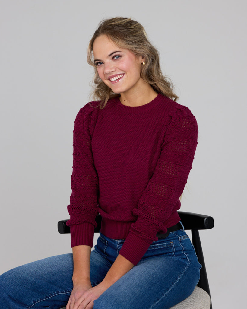 Model wearing a burgundy long sleeve sweater with pom pom details down sleeves.