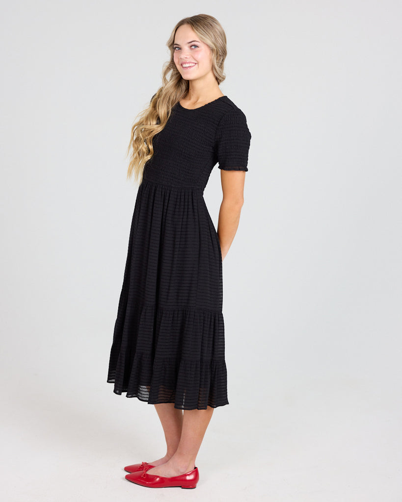 Model wearing a black short sleeve, round neck, midi length dress with a smocked bodice.