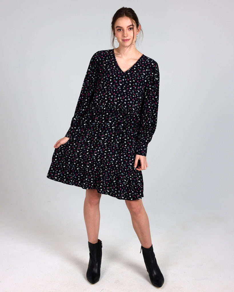 Model wearing a black dress with grey, white and red dainty floral print, with long sleeves, a v-neck and tiers that is knee length.