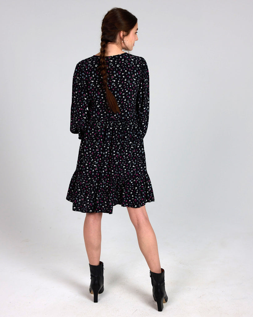 Model wearing a black dress with grey, white and red dainty floral print, with long sleeves, a v-neck and tiers that is knee length.