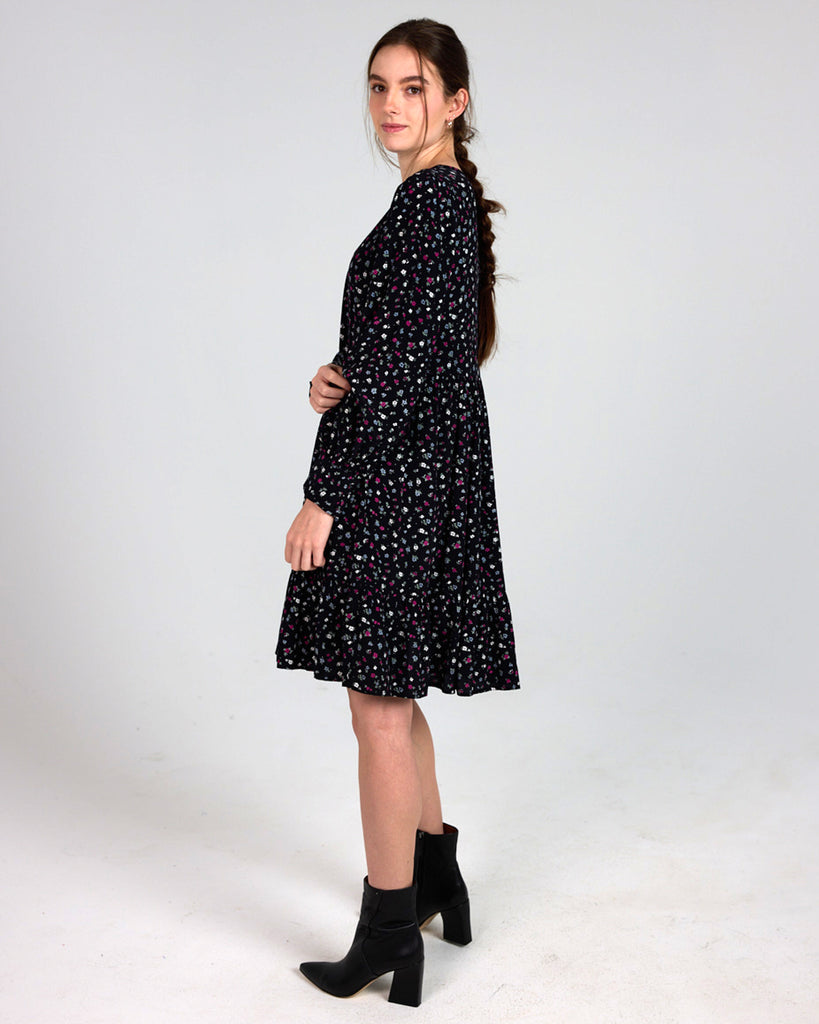 Model wearing a black dress with grey, white and red dainty floral print, with long sleeves, a v-neck and tiers that is knee length.