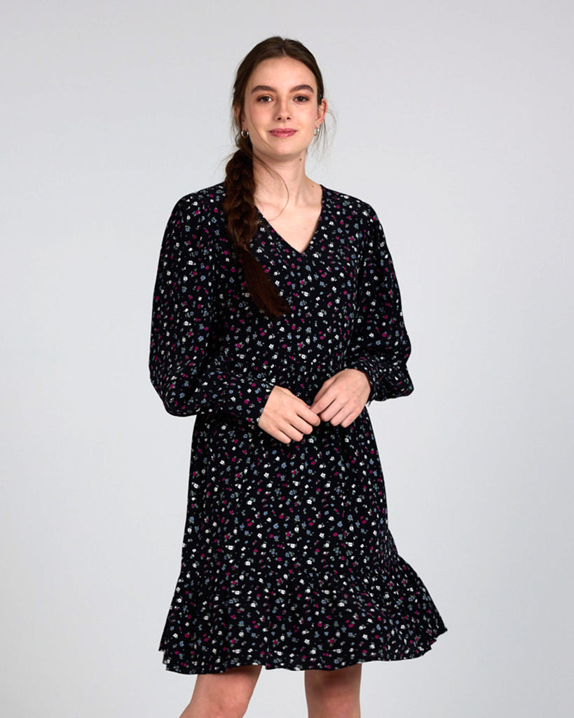 Model wearing a black dress with grey, white and red dainty floral print, with long sleeves, a v-neck and tiers that is knee length.