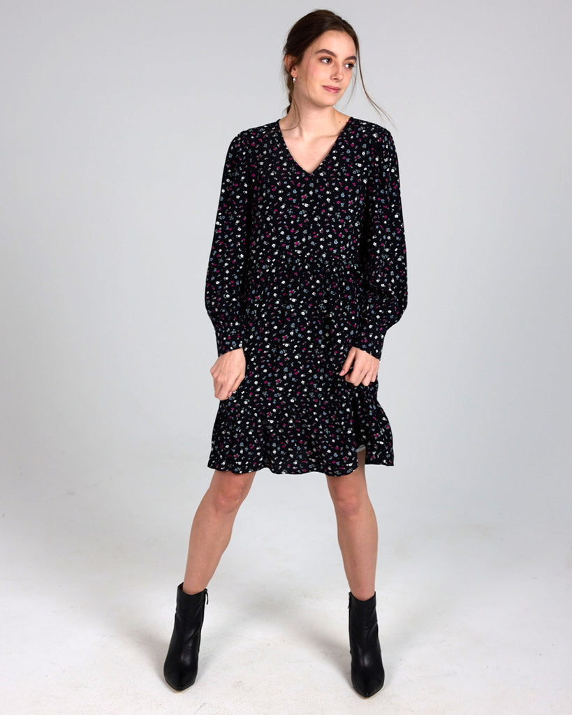 Model wearing a black dress with grey, white and red dainty floral print, with long sleeves, a v-neck and tiers that is knee length.