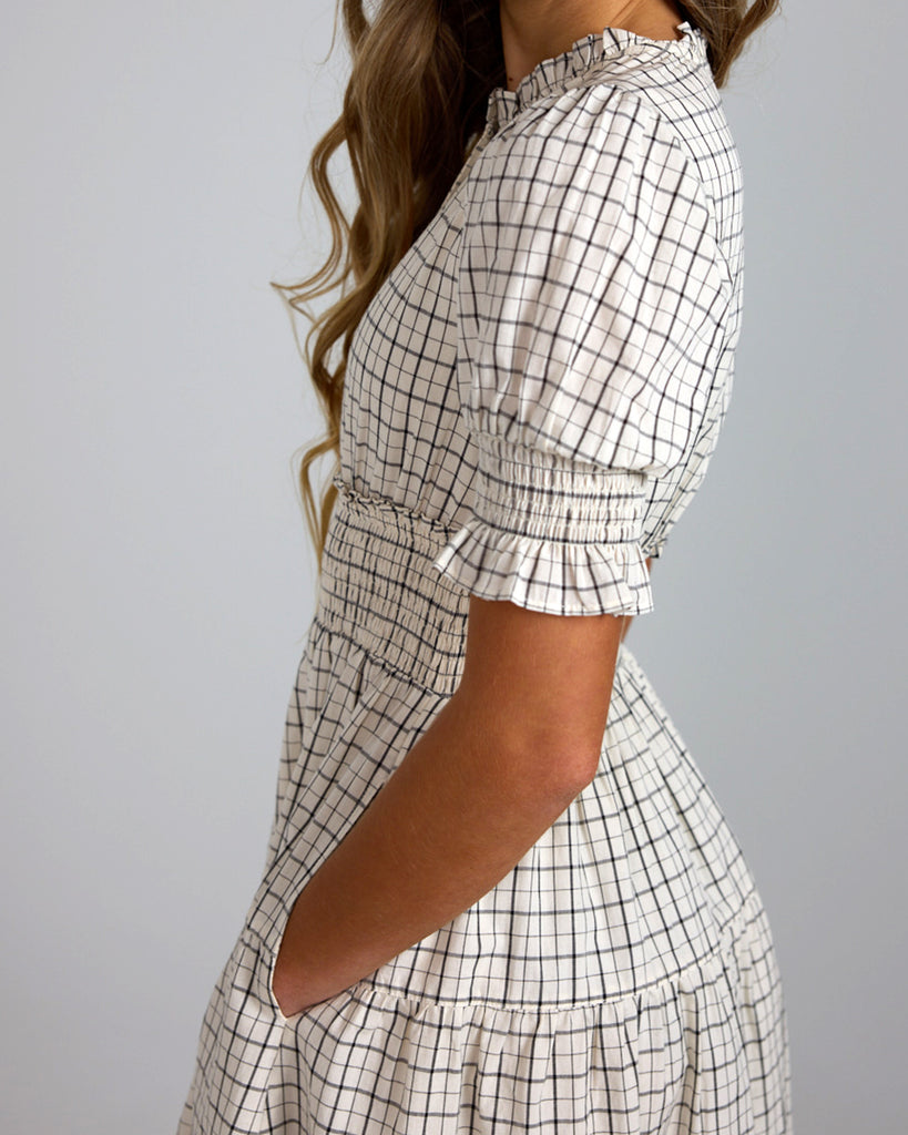 Up close shot of model wearing a white and grey plaid short sleeve mock neck midi-length dress with smocking details around waist.