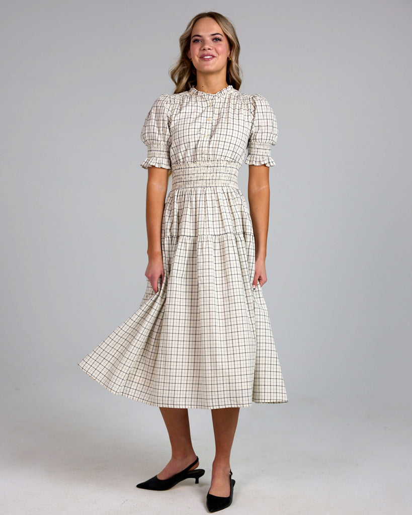 Model wearing a white and grey plaid short sleeve mock neck midi-length dress with smocking details around waist.