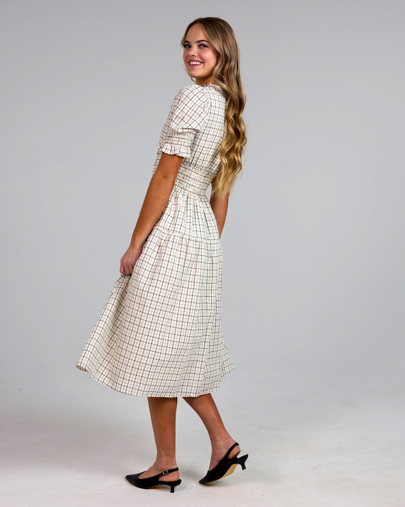 Model wearing a white and grey plaid short sleeve mock neck midi-length dress with smocking details around waist.