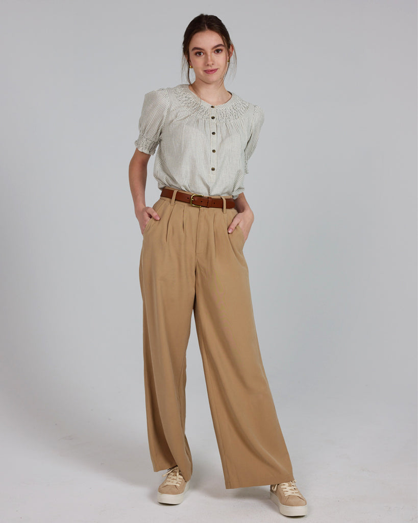 Model wearing a beige short sleeve blouse with button details and a small plaid pattern with tan trousers.