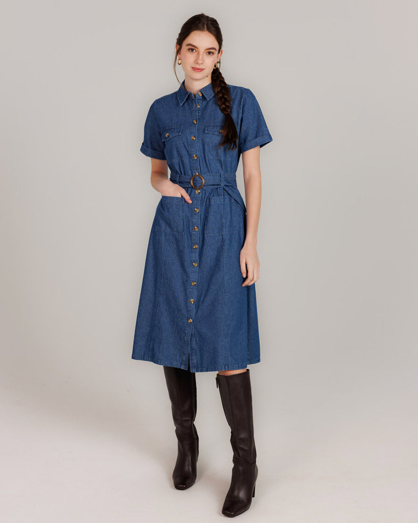 Model wearing a blue denim short sleeve, collared, knee-length dress with buttons down the front and a sash around the waist.