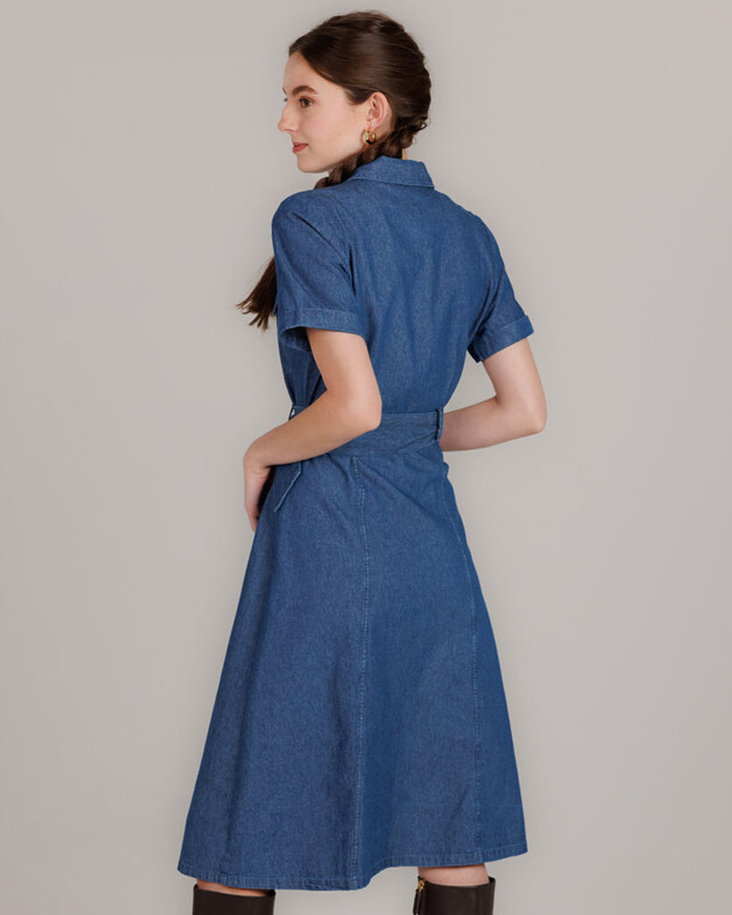 Model wearing a blue denim short sleeve, collared, knee-length dress with buttons down the front and a sash around the waist.