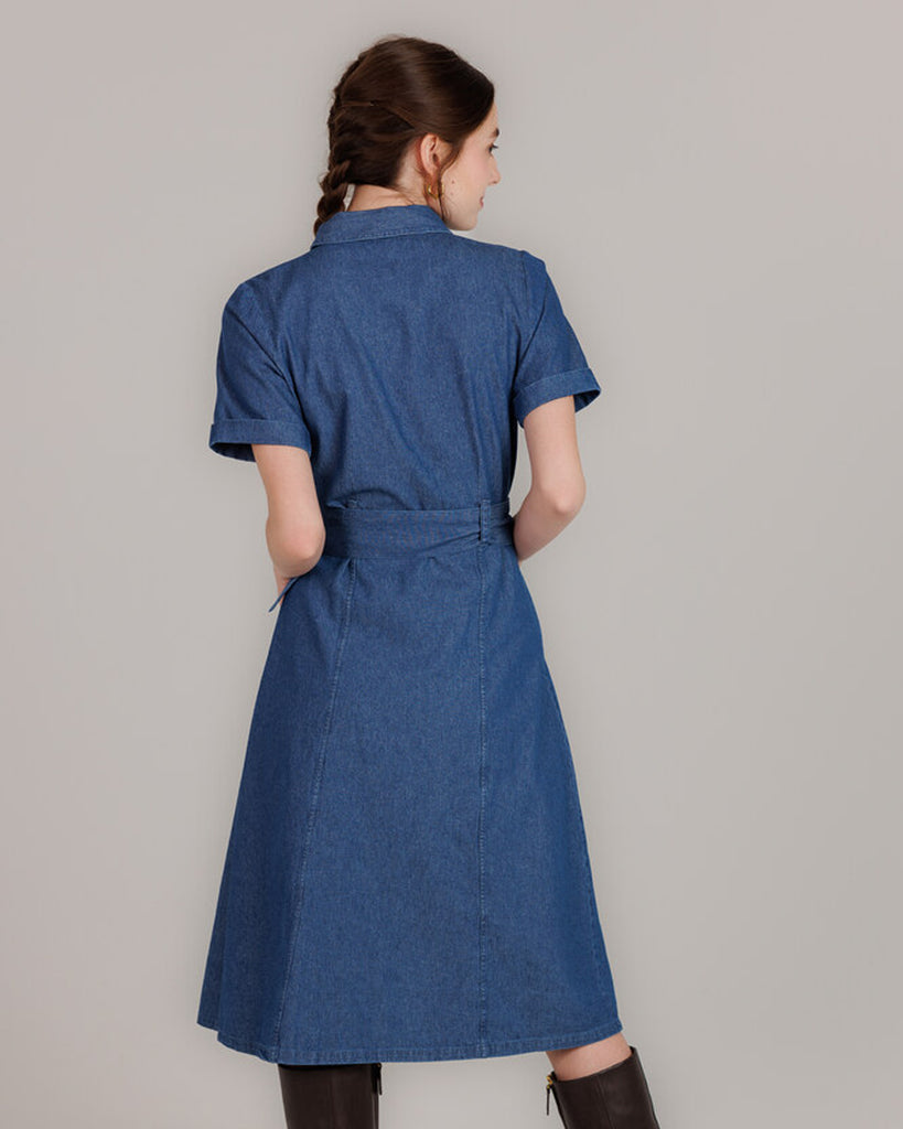 Model wearing a blue denim short sleeve, collared, knee-length dress with buttons down the front and a sash around the waist.