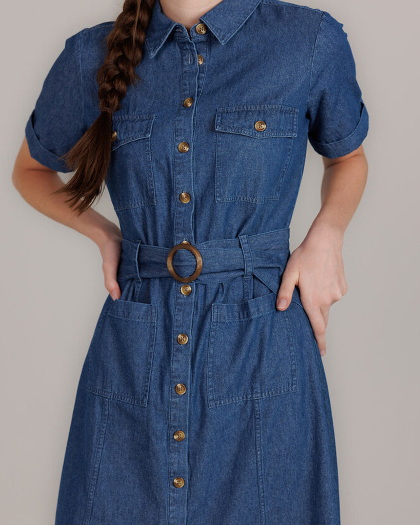 Model wearing a blue denim short sleeve, collared, knee-length dress with buttons down the front and a sash around the waist.