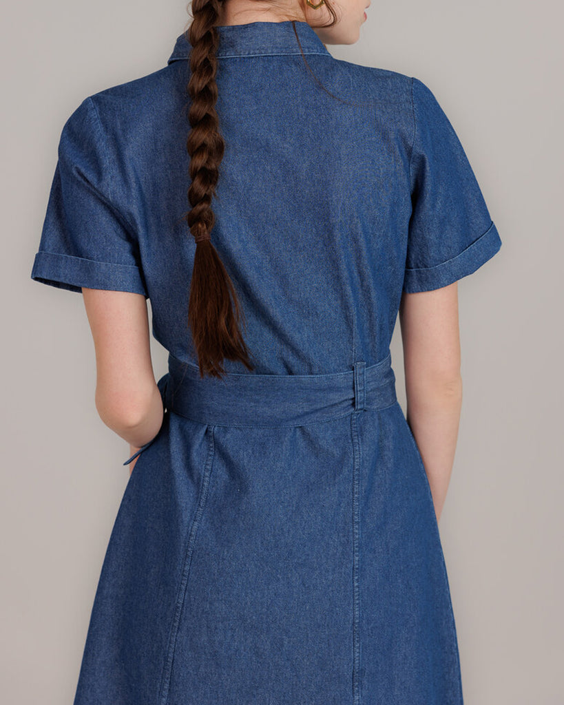 Model wearing a blue denim short sleeve, collared, knee-length dress with buttons down the front and a sash around the waist.